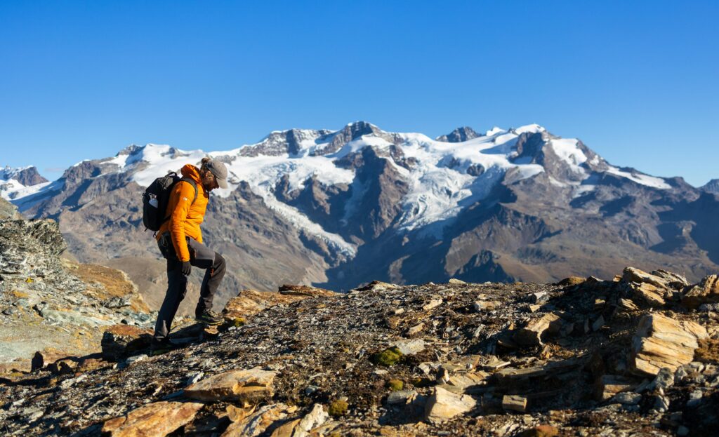 Solo hiker trekking through rocky alpine terrain with snow-capped Swiss Alps in the background under clear blue skies.