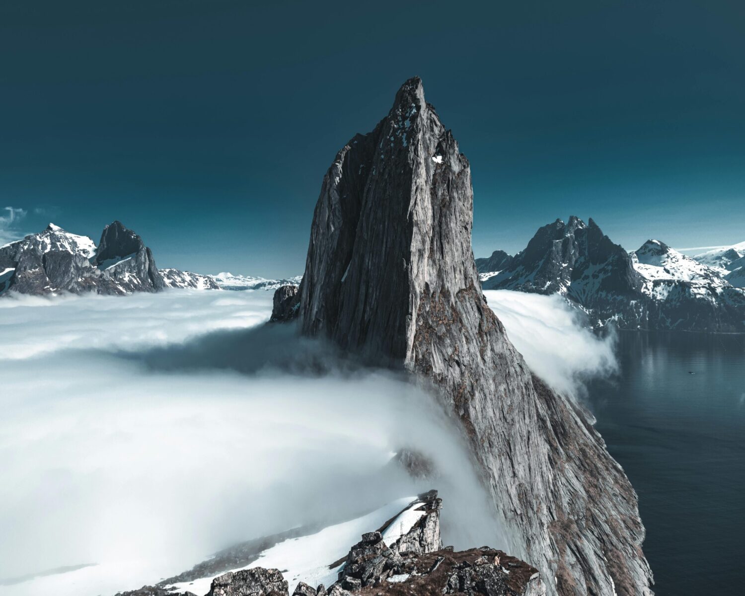 Towering jagged mountain peak rising above clouds with snowy Norwegian fjords in the distance