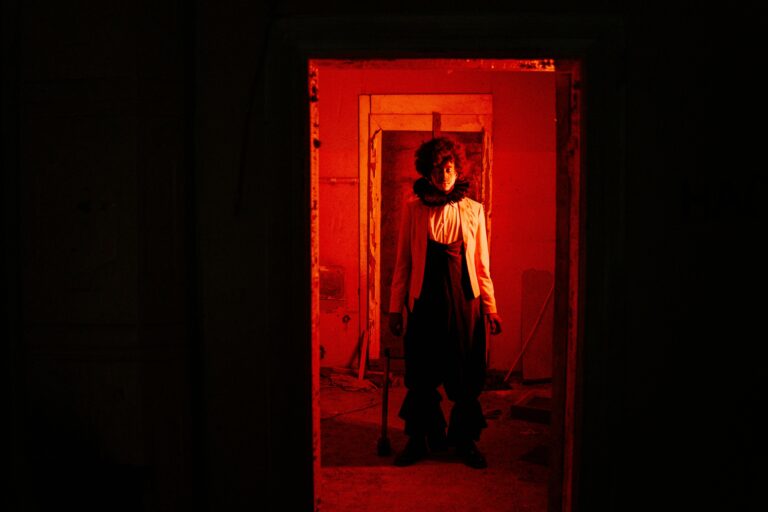 A mysterious figure in dramatic costume stands in a red-lit room, creating a tense and unsettling atmosphere.