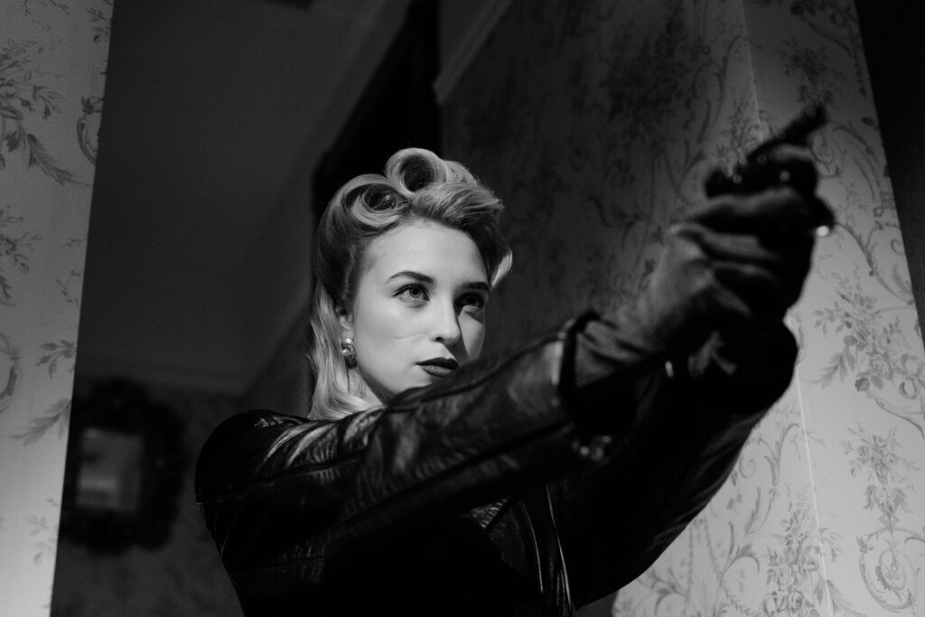 “Black-and-white shot of a woman with vintage waves, clad in a leather jacket and gloves, aiming a pistol down a narrow, patterned hallway—evoking classic film-noir suspense.”