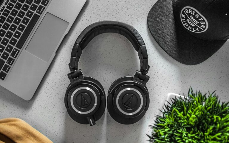 “Overhead shot of black Audio-Technica over-ear headphones resting on a speckled gray desk beside a silver laptop keyboard, a black Brixton cap, and a small potted green plant.”