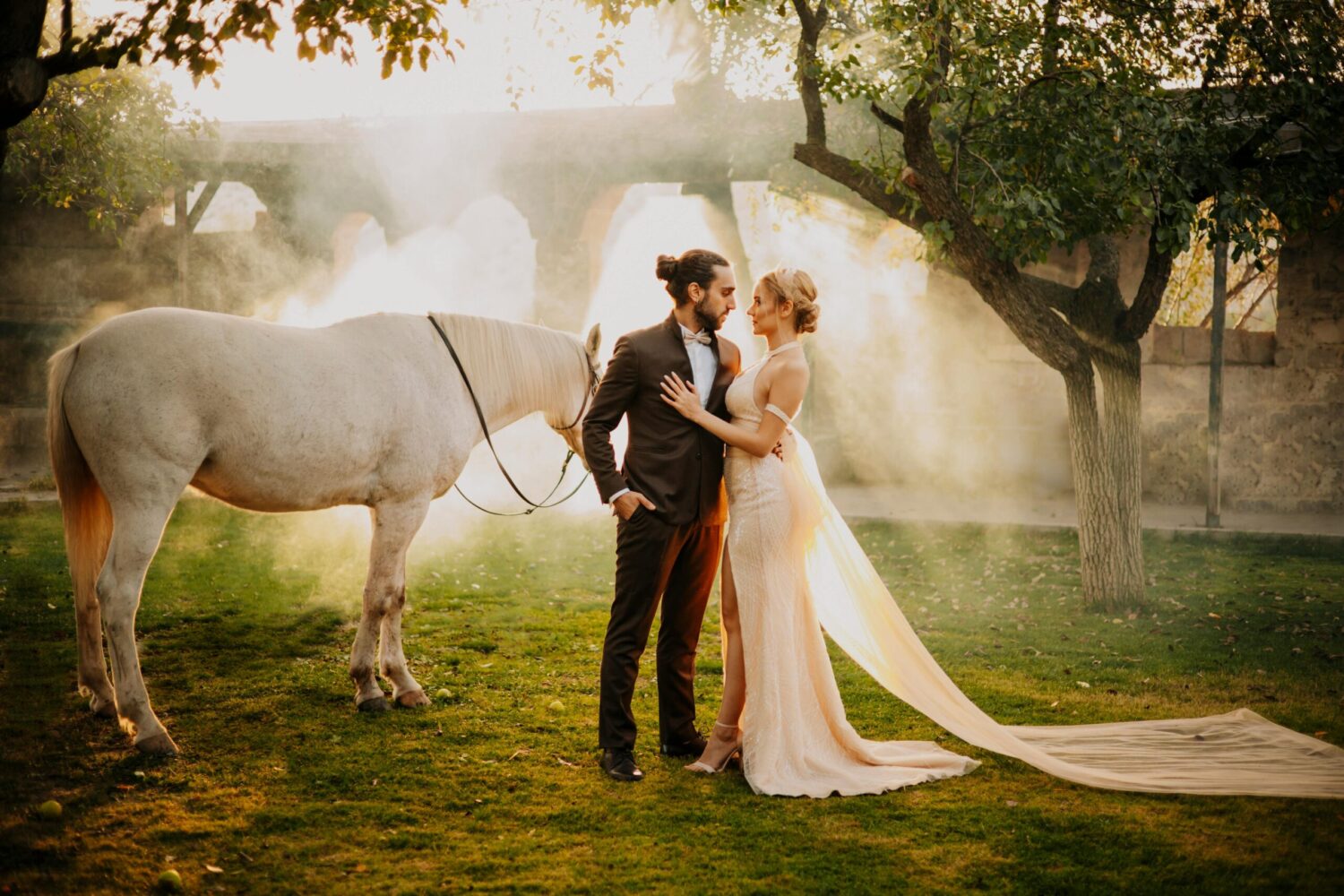A romantic couple in wedding attire stands in a sunlit garden with a white horse beside them, surrounded by mist and trees.